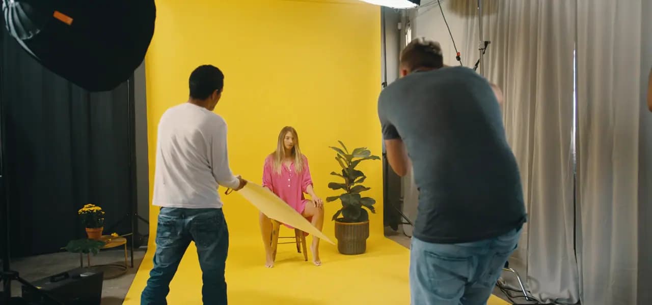A studio shoot on a yellow backdrop: a photographer frames a seated model while an assistant angles a reflector.